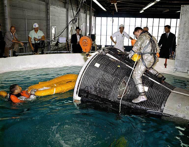 stronauts John Young and Virgil I. (Gus) Grissom are pictured during water egress training in a large indoor pool at Ellington Air Force Base, Texas. Young is seated on top of the Gemini capsule while Grissom is in the water with a life raft.
